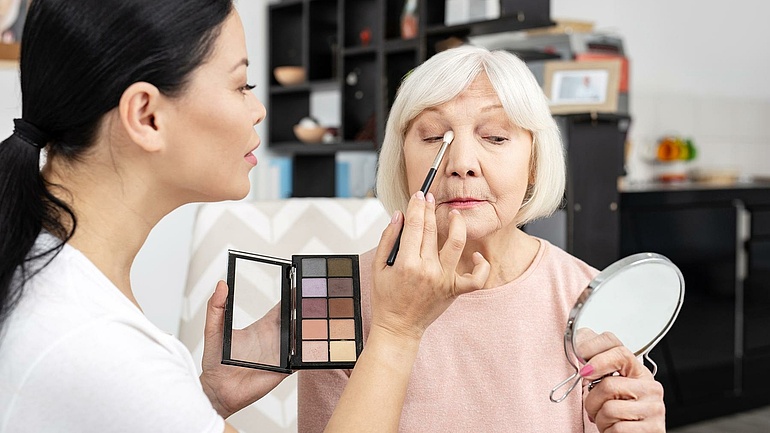 Eine junke Kosmetiker schminkt eine ältere Dame, die sich währenddessen in einem Handspiegel betrachtet.