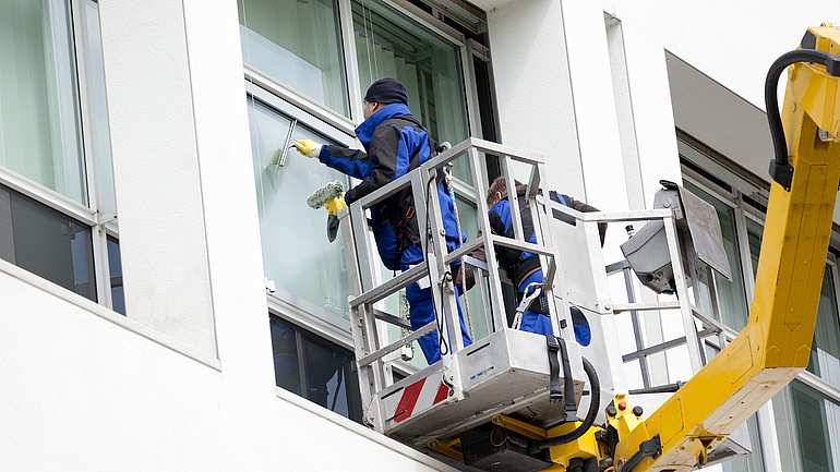 Gebäudereiniger stehen in einer Hubarbeitsbühne und putzen Fenster.