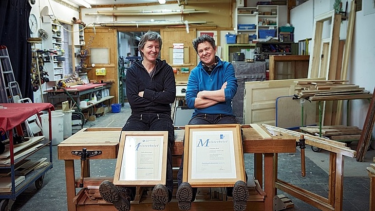 Zwei Tischlerinnen mit eingerahmten Meisterbrief in ihrer Werkstatt.